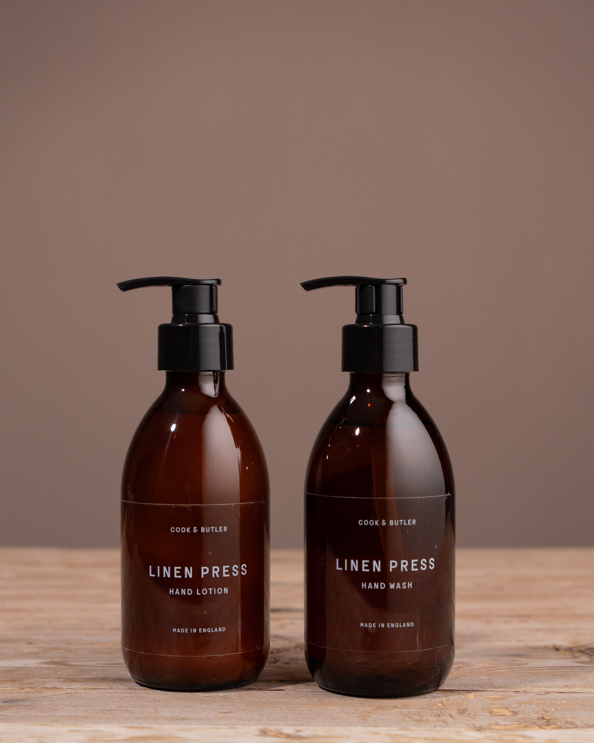 Two bottles of 'Linen Press' Cook & Butler hand care products on a wooden surface with a brown background.