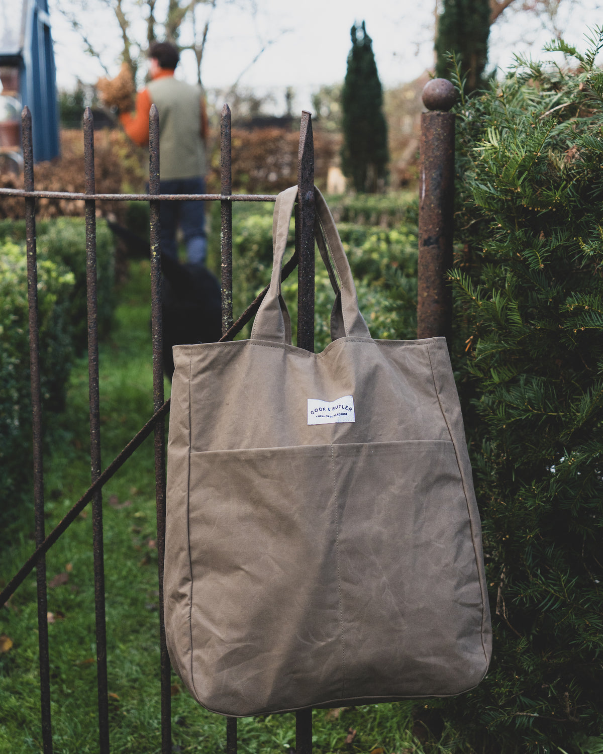 Brown tote bag with a Cook & Butler brand label hanging on a metal gate outdoors.