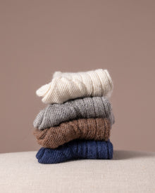 Stack of four pairs of socks in white, grey, brown, and blue on a beige surface with a beige background.