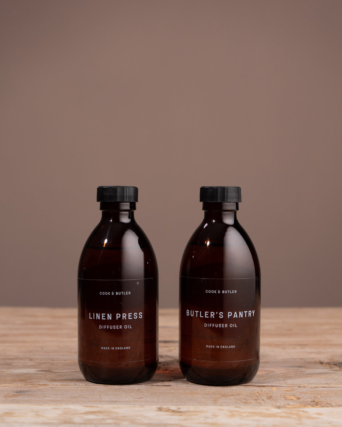 Two bottles of Cook & Butler diffuser oil 'Butler's Pantry' on a wooden surface with a brown background.