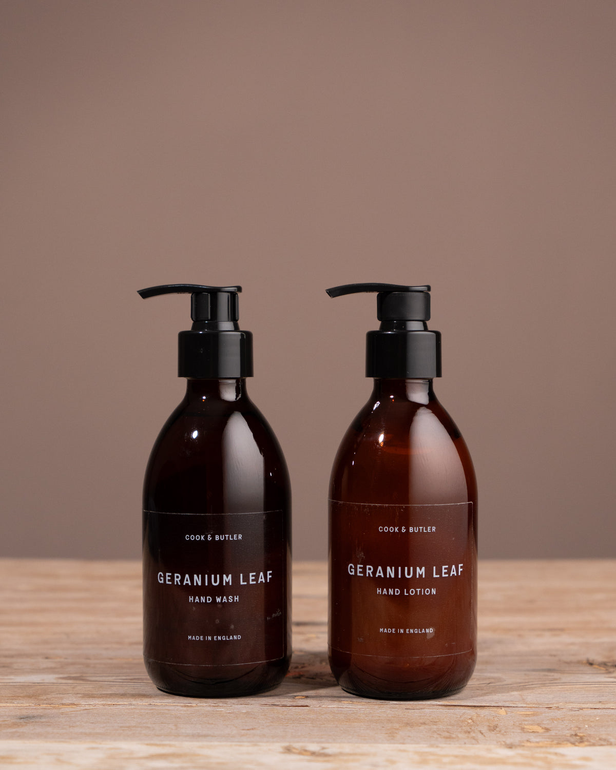 Two bottles of Cook & Butler 'Geranium Leaf' hand wash and lotion on a wooden surface with a brown background.