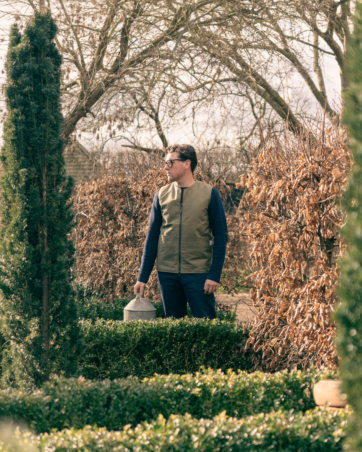 Man wearing a green field vest standing in a garden with trees and shrubs