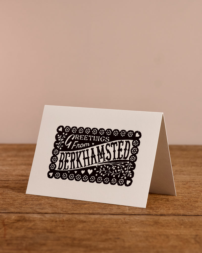 Greeting card with 'Greetings from Berkhamsted' text on a wooden surface and beige background