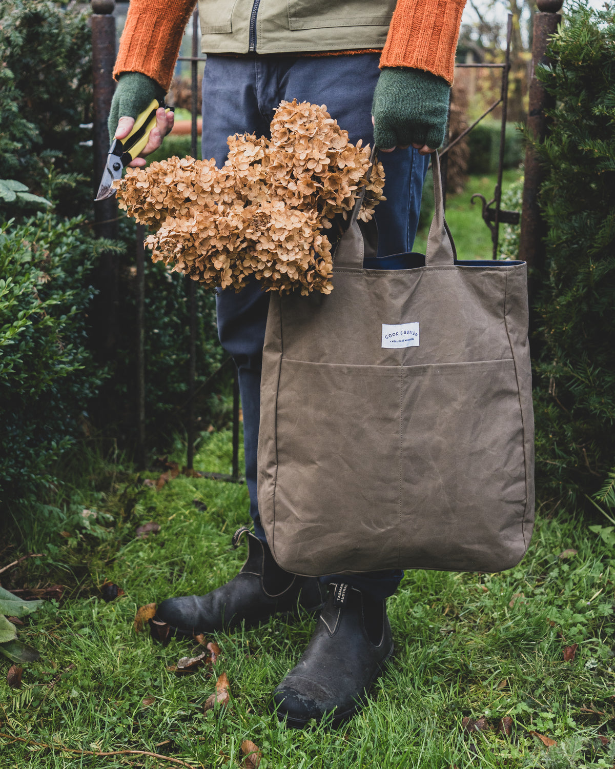 Person holding a brown tote bag filled with dried plants in a garden setting
