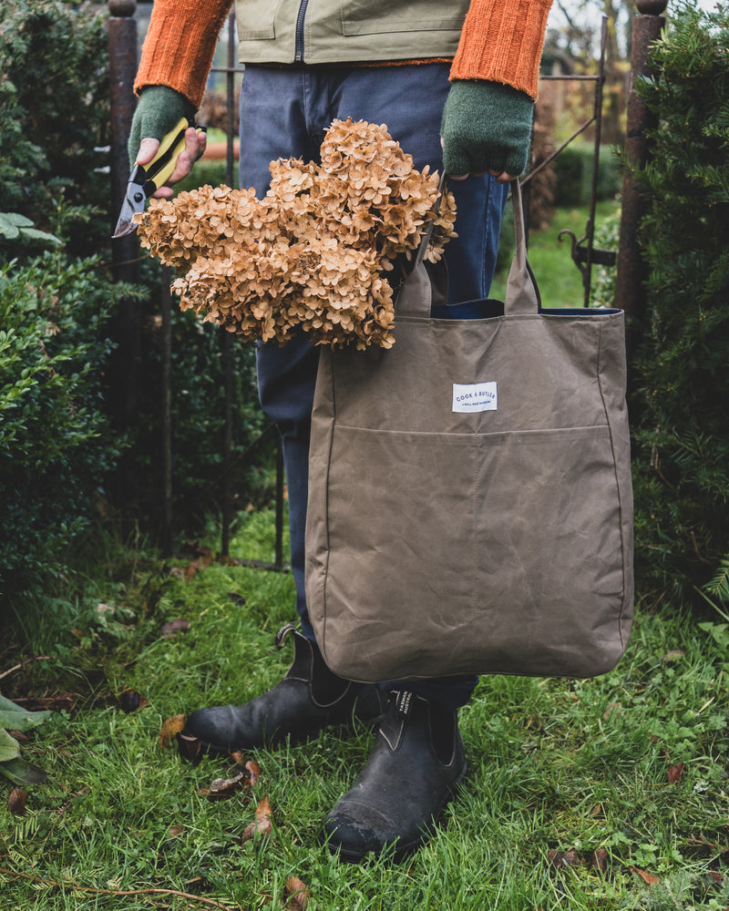 Person holding a brown tote bag filled with dried plants in a garden setting