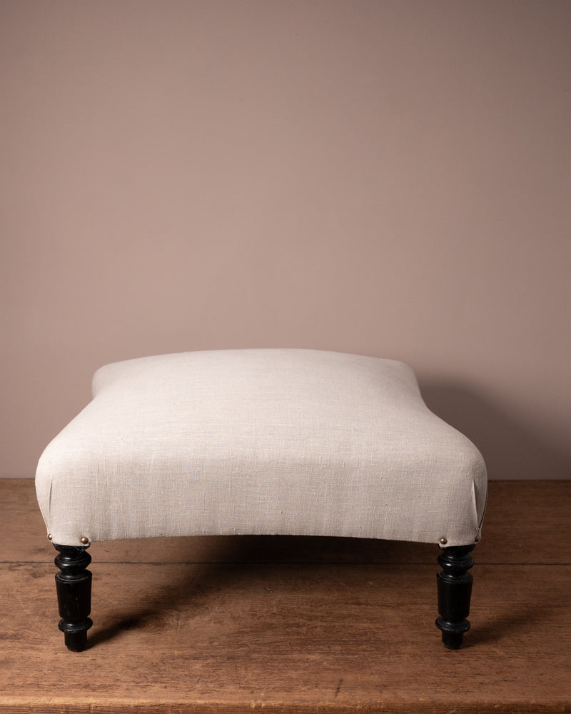White upholstered ottoman with black legs on a wooden floor against a beige wall
