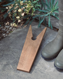 Wooden boot jack on a concrete surface with green boots and plants in the background