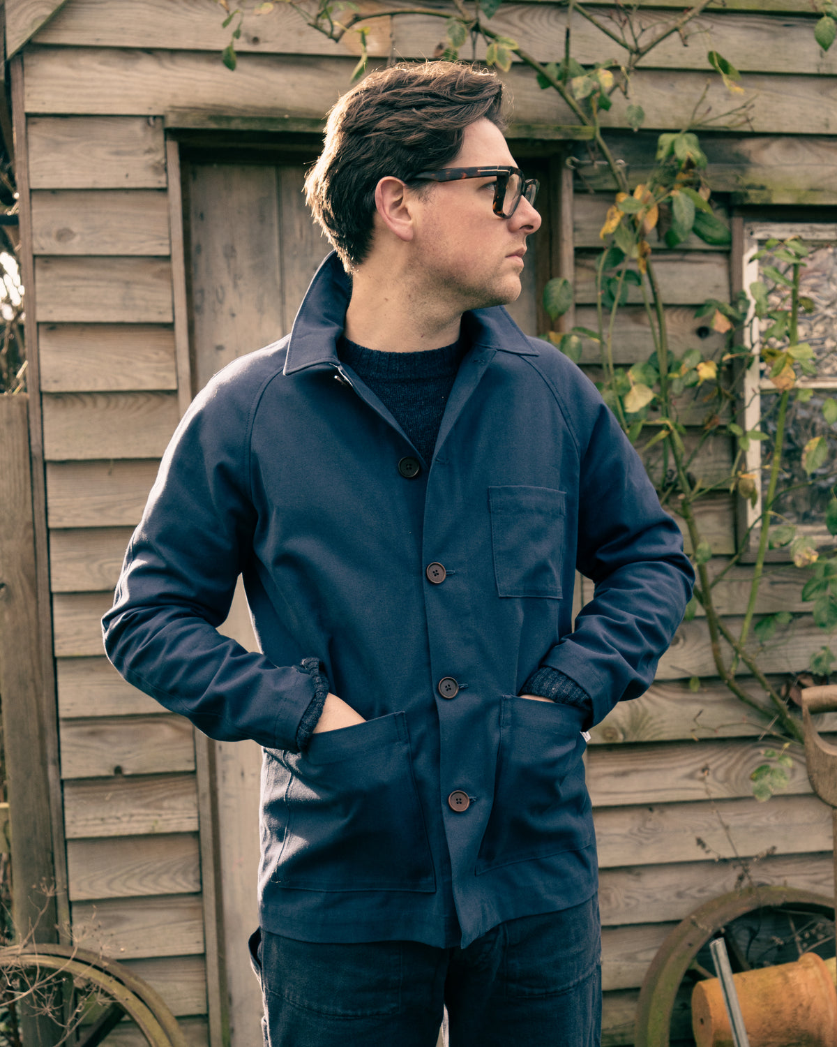 Man wearing a blue Cook & Butler work jacket standing in front of a wooden shed.