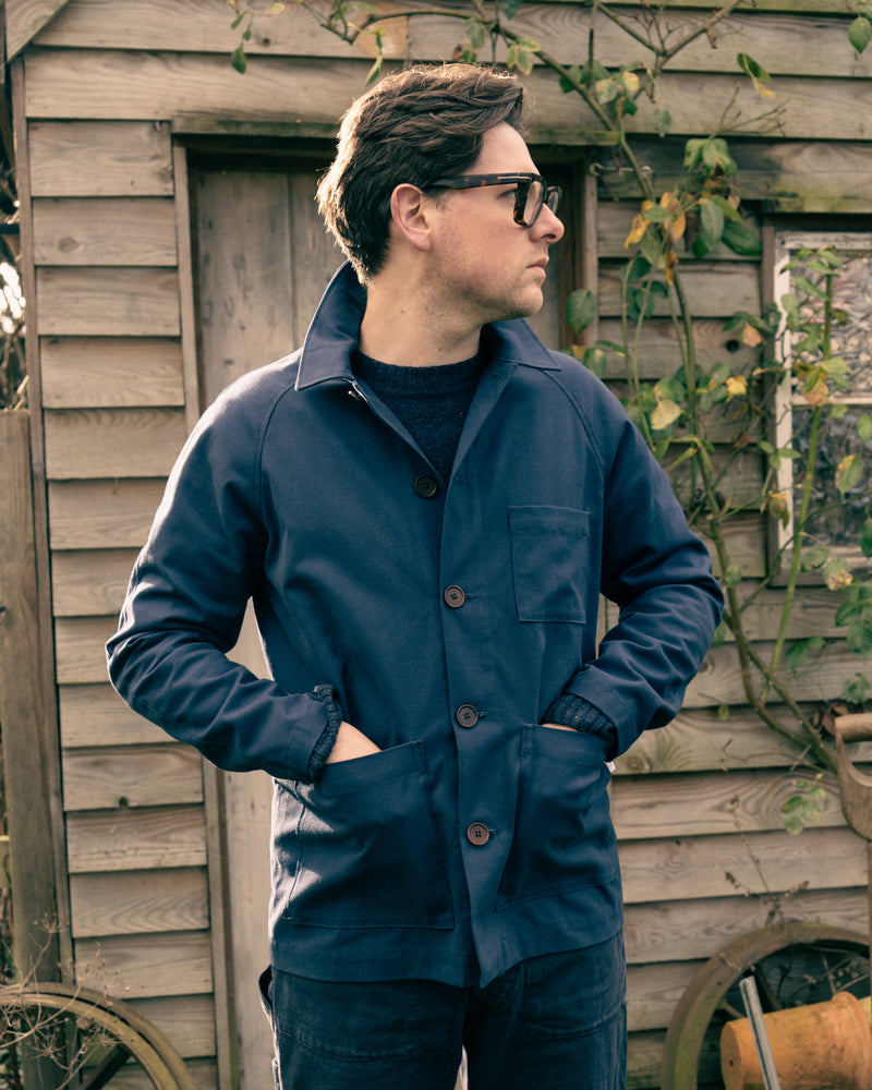 Man wearing a blue Cook & Butler work jacket standing in front of a wooden shed.